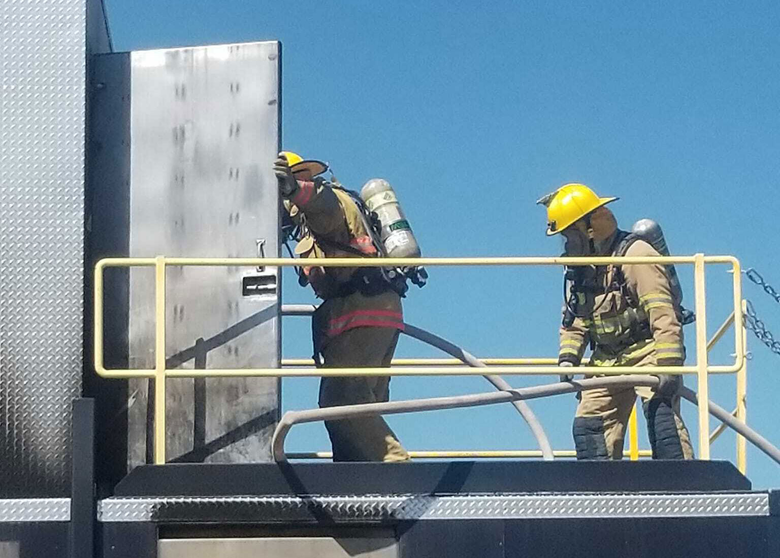 Two firefighters in full gear, including helmets and oxygen tanks, stand on a metal platform near a tall, metallic structure. One is opening a heavy door while holding a hose, and the other stands behind, assisting. Blue sky in the background.